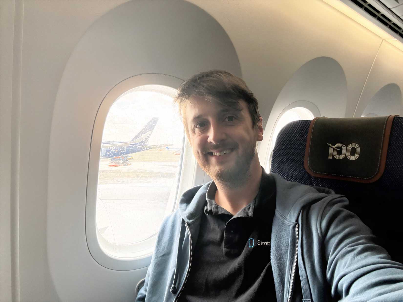 A smiling man sits in an aircraft window seat, wearing a blue zip-up hoodie over a black shirt with a "Simple Flying" logo. The headrest of his navy blue seat features a silver "100" commemorative logo. Through the window, another aircraft with a blue and white tail is visible on a sunny tarmac.