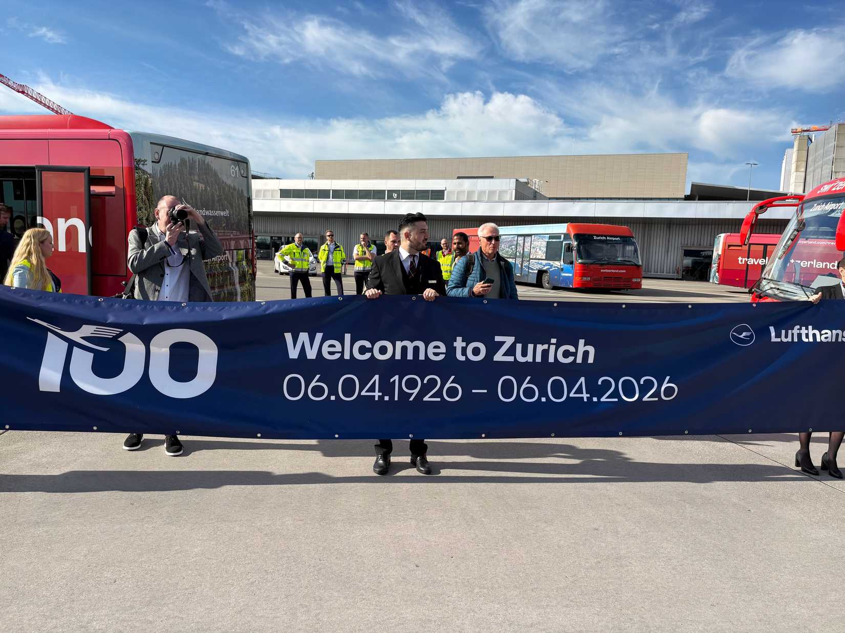 Several people hold a large navy blue banner on an airport apron that reads "Welcome to Zurich" and "06.04.1926 – 06.04.2026." The banner features the Lufthansa logo and a commemorative "100" graphic, while red airport buses and various staff members are visible in the background under a bright, partly cloudy sky.