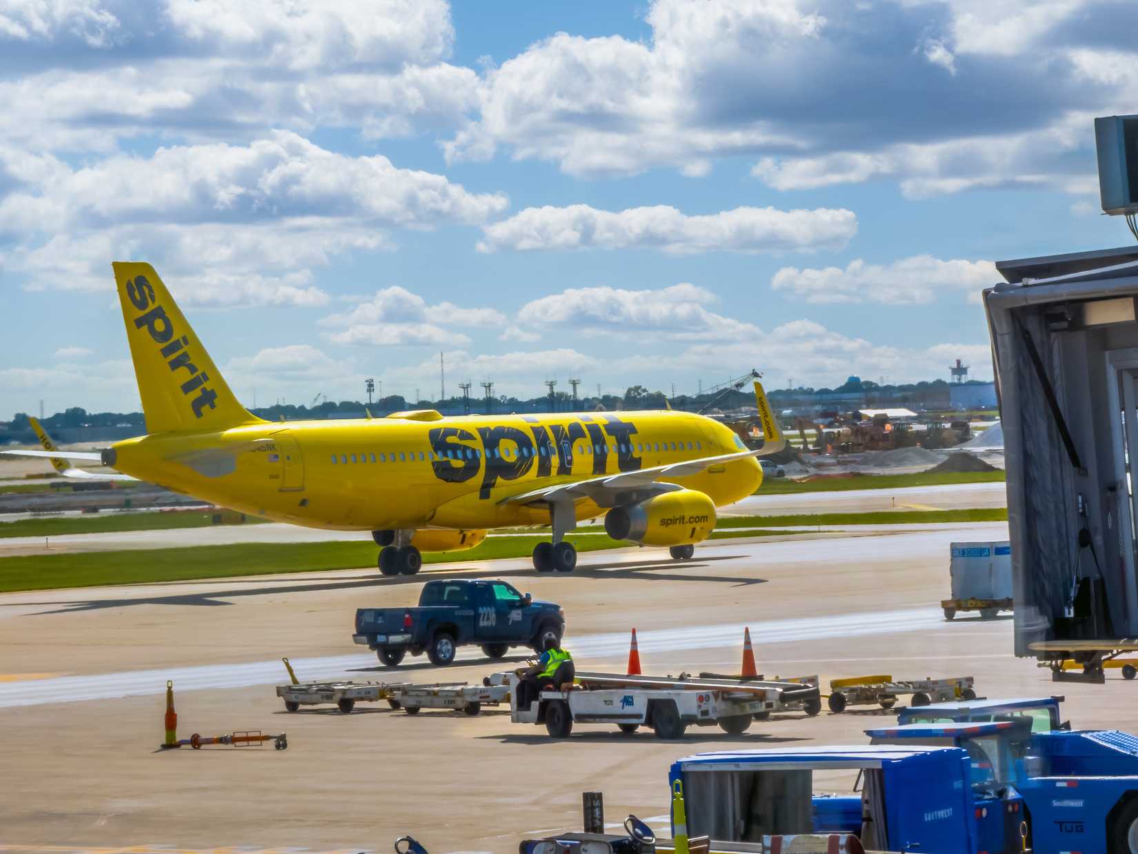 A yellow Spirit Airlines jet taxis on the tarmac past ground crew and service vehicles under a cloudy sky.
