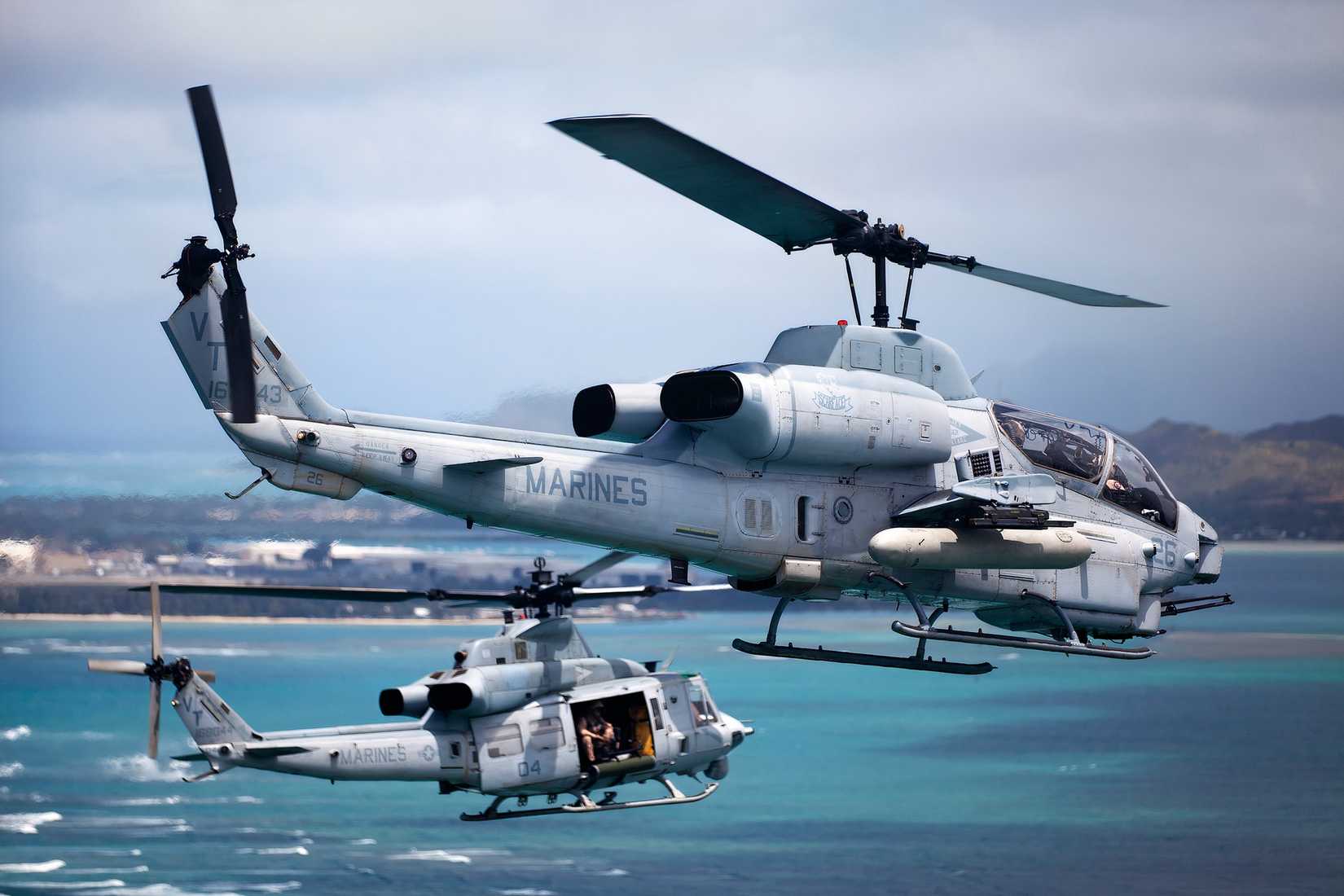 A Marine Corps AH-1 Cobra flying with another helicopter towards over the water towards an island.