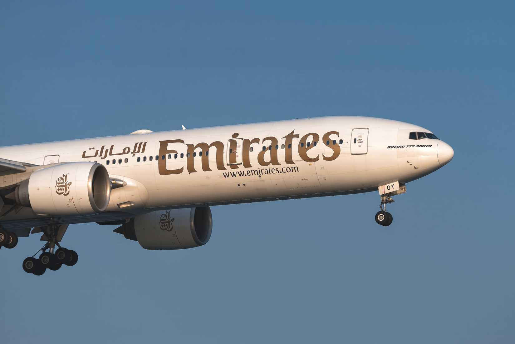 Emirates Boeing 777-300ER passenger aircraft flying with landing gear deployed under blue sky.