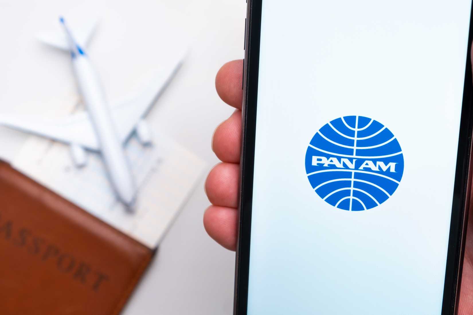 Pan Am Airline logo on the mobile phone screen with a plane, passport and boarding pass on the background. The concept of the airlines mobile application. 