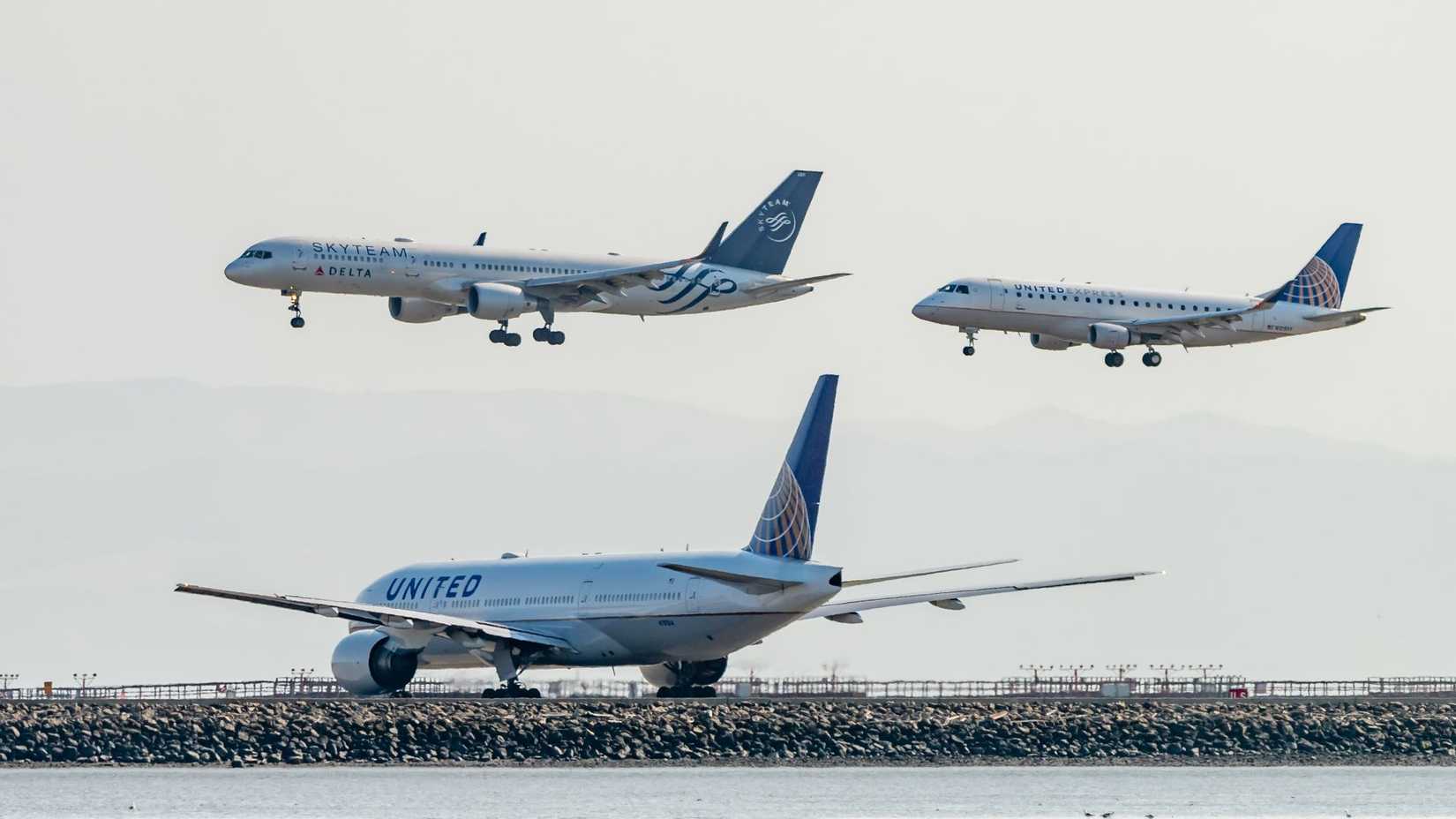 A United Airlines plane and Delta Airlines (with SkyTeam livery) plane come into land while a United Airlines plane waits at the end of the runway, 2018.