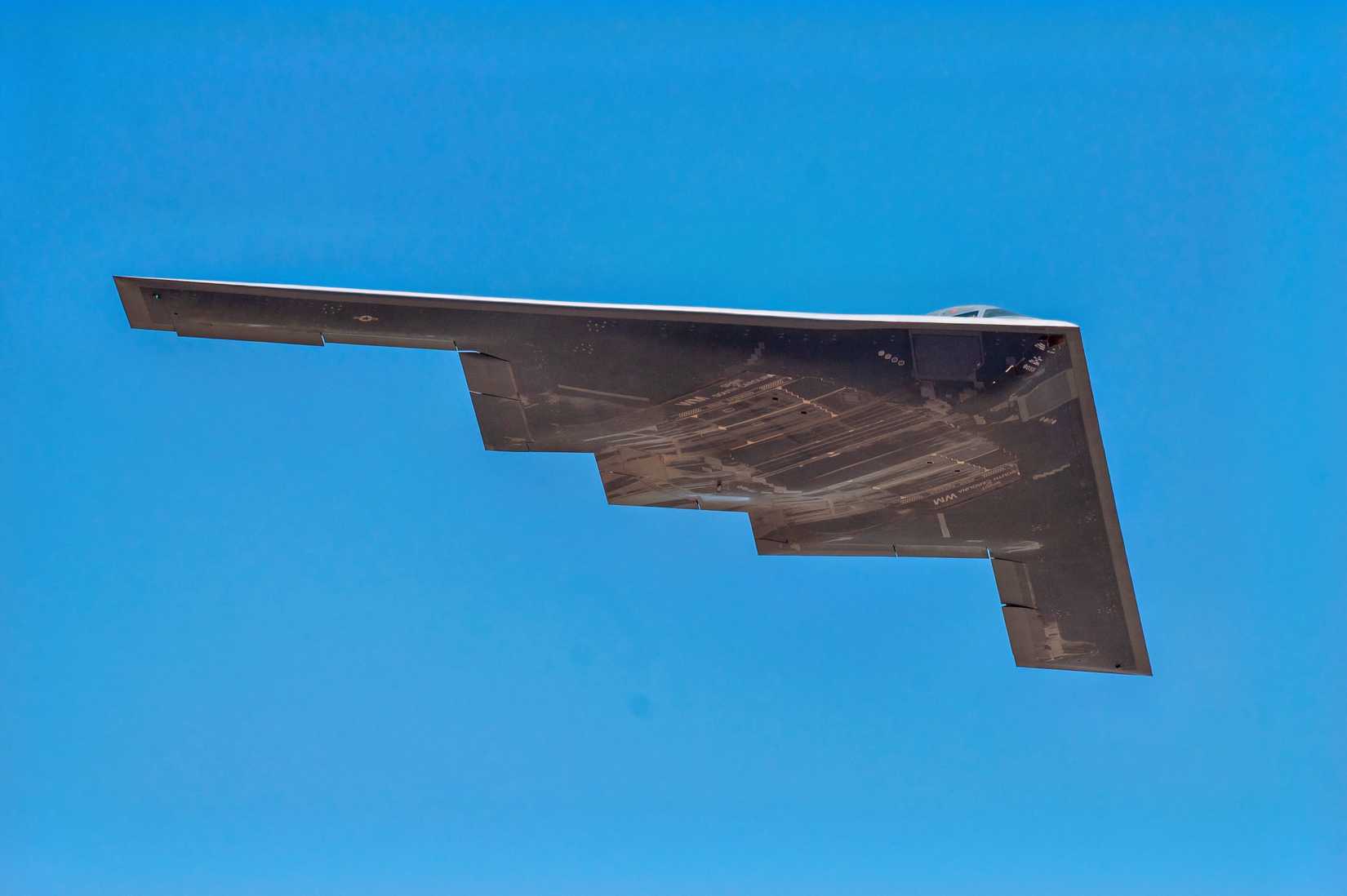A photo of the underside of a B-21 flying against backdrop of a blue sky.
