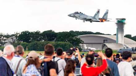 A crowd of spectators looks on and takes photos from behind a barrier as an F-15 fighter jet takes off with glowing orange afterburners, climbing low over airport hangars and an air traffic control tower against an overcast sky.