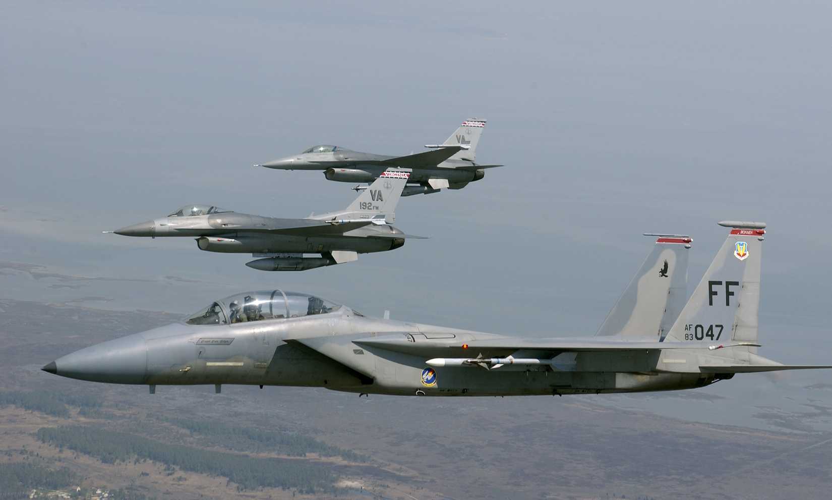Two F-16s and an F-15 flying in a tight formation over a rural area, against a gray sky.