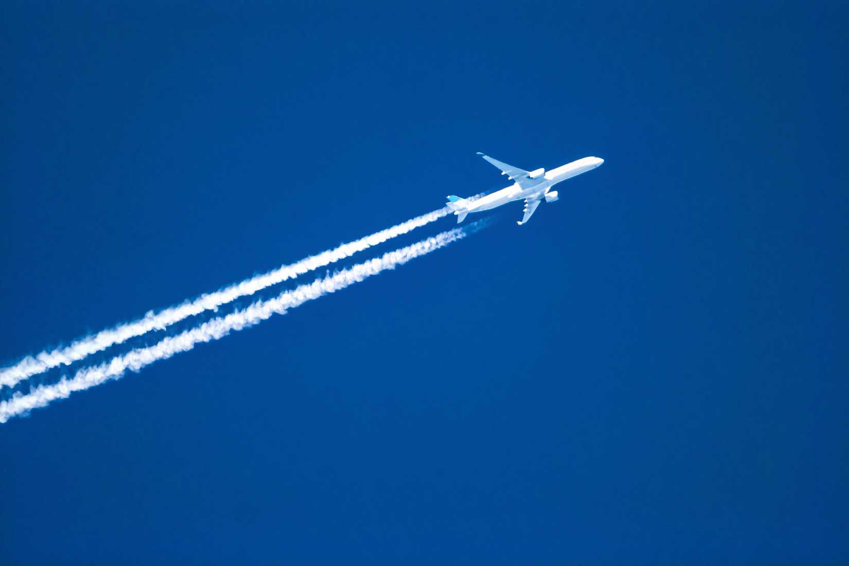  A white commercial passenger jet flying at high altitude, angled toward the upper right against a solid, deep blue sky, leaving two prominent parallel white vapor contrails in its wake.