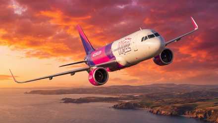 A Wizz Air commercial airliner flying high above a scenic, rugged coastline during a vibrant, fiery orange and pink sunset that fills the sky with dramatic clouds.