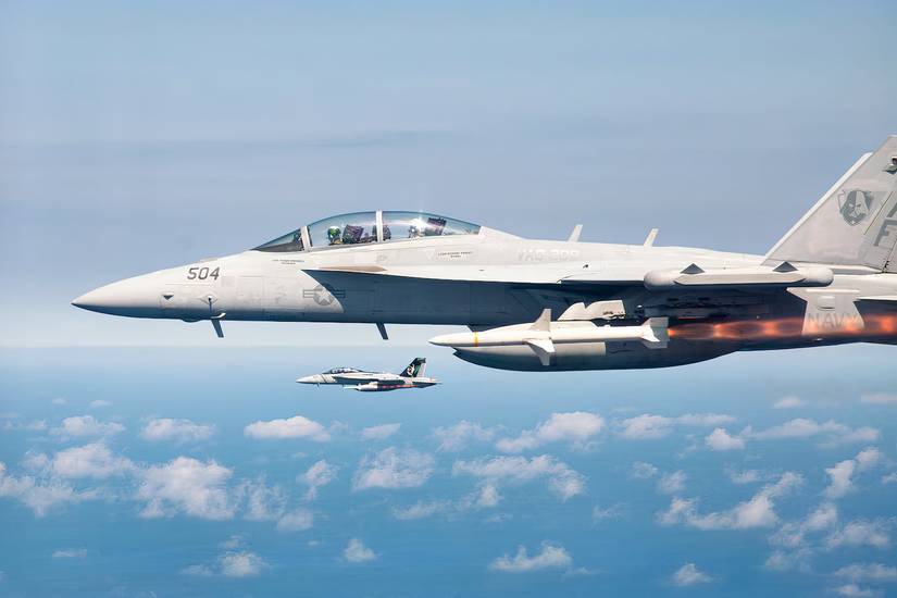 A photo of an FA-18 firing a missile with another F/A-18 in the background with light clouds and the sea below.