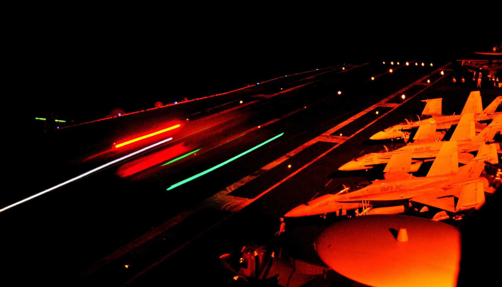 While one FA-18 Super Hornet takes off at night while three others are parked, presumably waiting their turn on an aircraft carrier deck. 