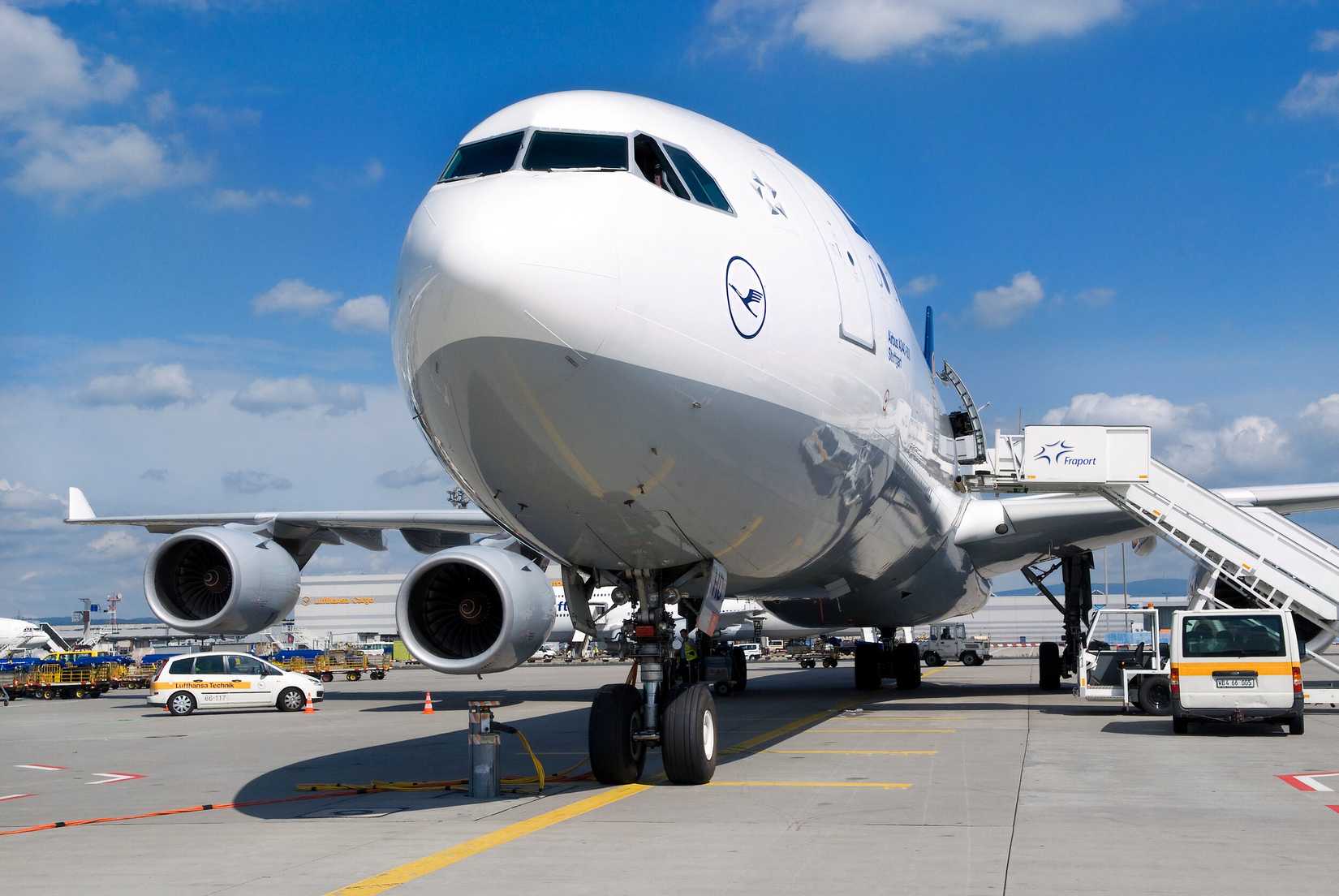 A low-angle, front-three-quarter view of a Lufthansa Airbus A340-600 parked on an airport apron. The massive widebody aircraft showcases its distinctive nose profile, cockpit windows, and two of its four engines on the port wing, while a Fraport mobile boarding stair truck is connected to the forward cabin door on the right side under a clear blue sky with scattered clouds.