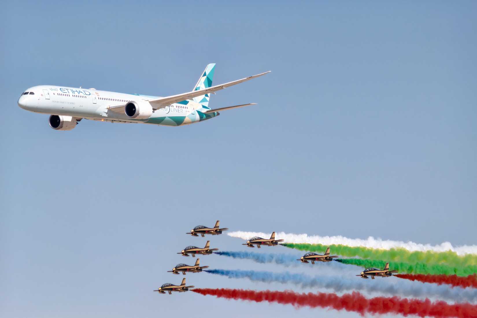 An Etihad Airways Boeing 787 Dreamliner flying in formation above the Al Fursan aerobatic display team. The seven jet aircraft fly closely below the airliner, trailing vibrant white, green, blue, and red smoke plumes against a clear blue sky.