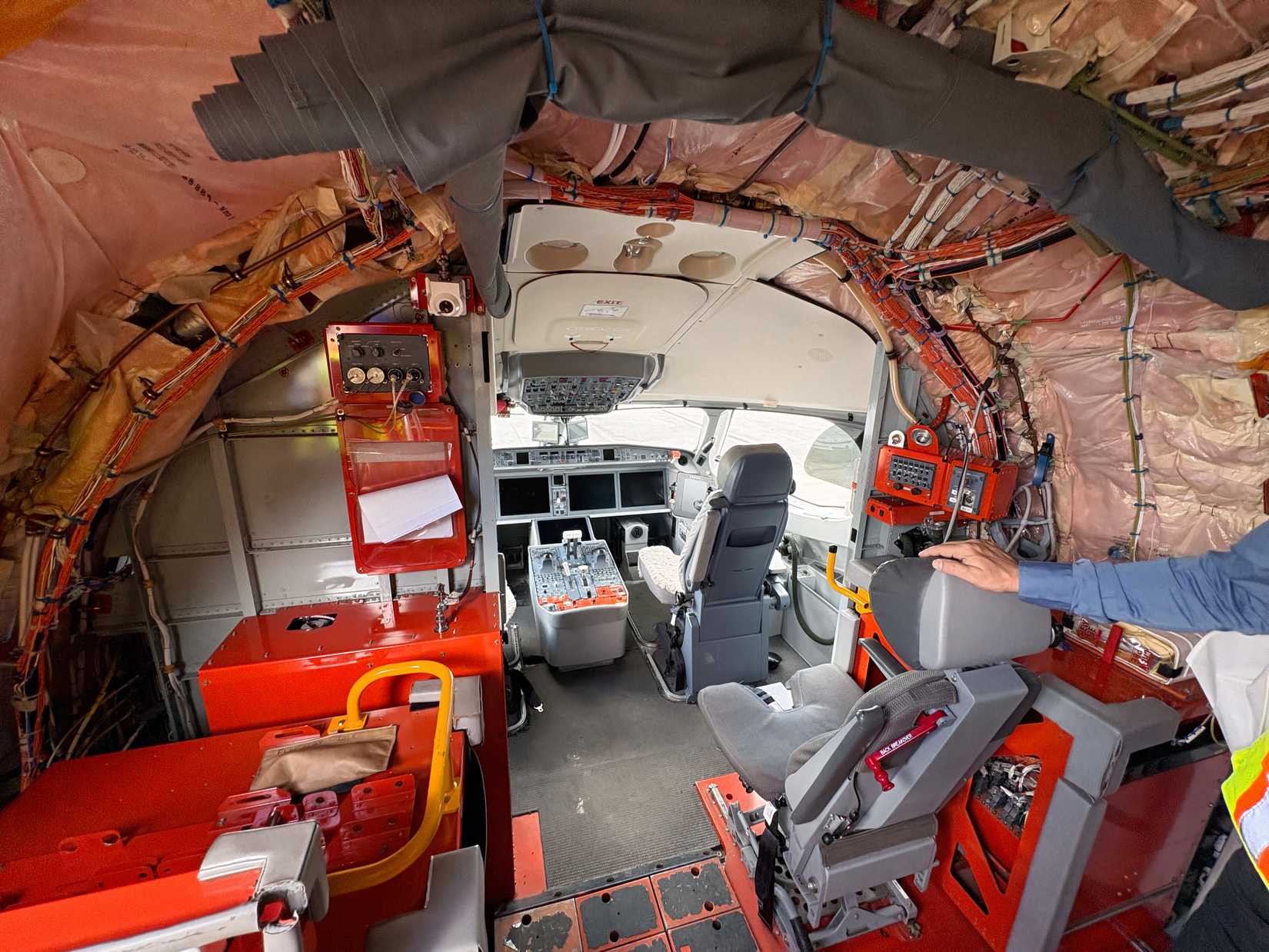 An interior viewpoint looking forward into an uncompleted aircraft cockpit, framed by exposed insulation padding and multi-colored wiring bundles on the fuselage walls. Bright red engineering equipment racks flank the entryway leading to the pilot seats and flight deck console.