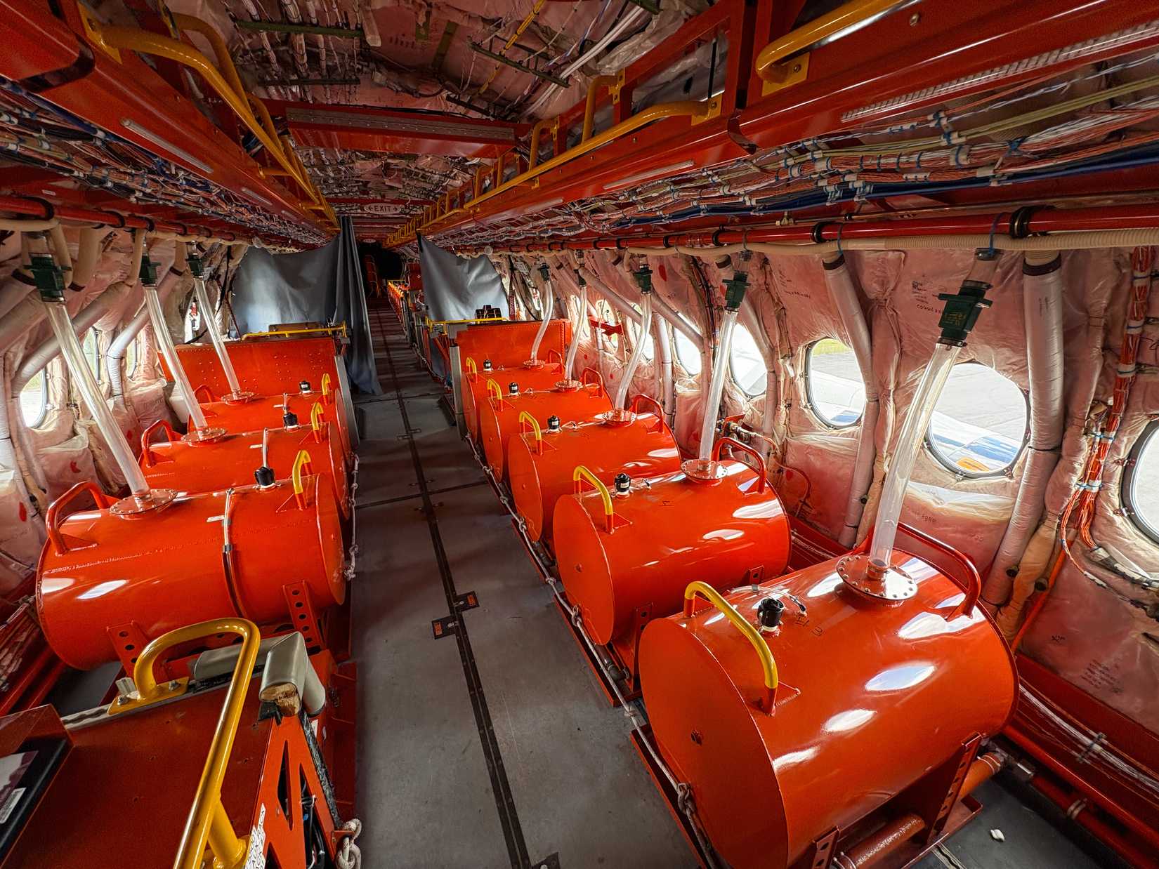 An interior view of an uncompleted test aircraft cabin, featuring rows of large, bright orange cylindrical ballast tanks instead of passenger seats. The exposed fuselage walls show insulation and extensive wiring bundles, with clear plastic tubes running from the tanks toward the upper structural framing along a central aisle.