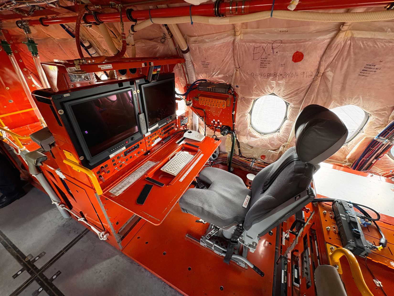 A flight-test engineer's workstation inside an uncompleted airplane cabin. The setup features a prominent bright orange desk assembly equipped with dual computer monitors, a keyboard, and an adjustable grey crew seat installed over exposed floor structures next to a cabin window.