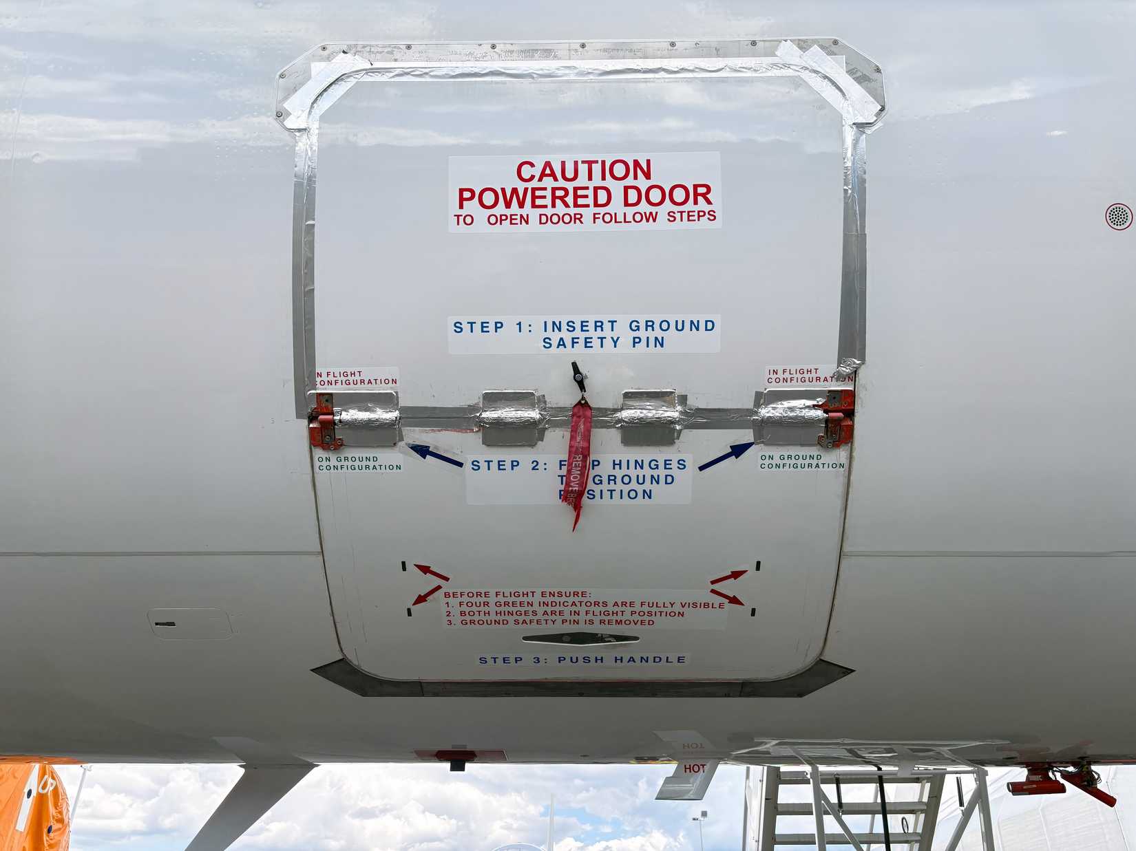 A close-up exterior view of an emergency door on a white test aircraft fuselage. The door features prominent red and blue printed operating instructions, metallic tape sealing the top edge, and a central hinge mechanism with a red warning streamer.