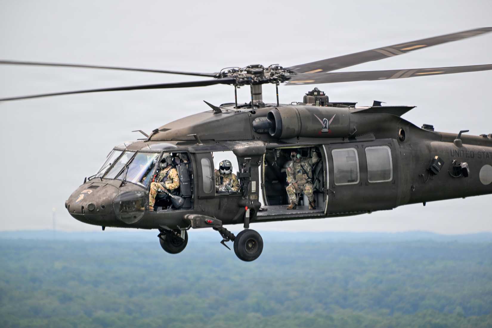 Army soldiers sit in the open cabin of a UH-60 Black Hawk helicopter during an airborne mission over dense forest