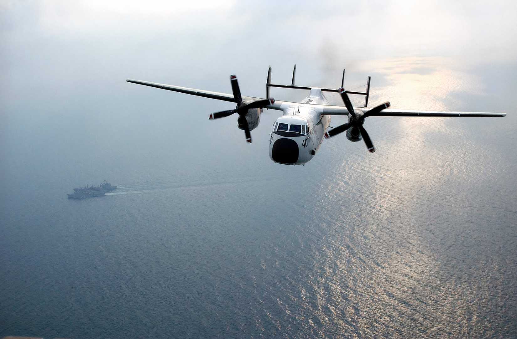 A photo of a Greyhound flying over the water with a carrier and a replenishment ship in the background. 
