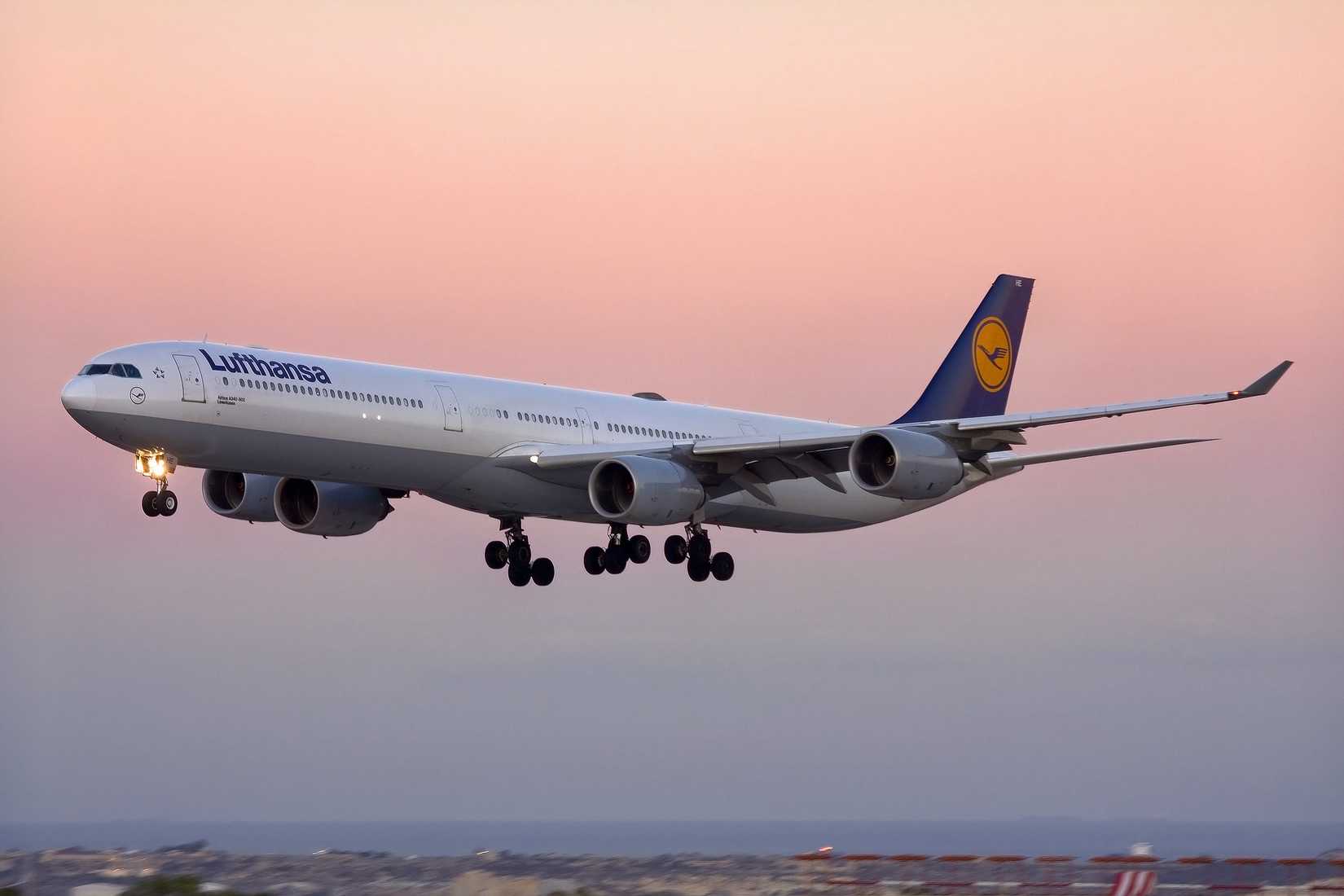 A large Lufthansa Airbus A340-600 commercial airliner captured on its final approach to landing. The long, four-engine widebody aircraft features the classic Lufthansa livery with a white fuselage, grey underside, and a dark blue tail fin boasting the golden crane logo, all set against a beautiful gradient pastel pink and orange sunset sky.