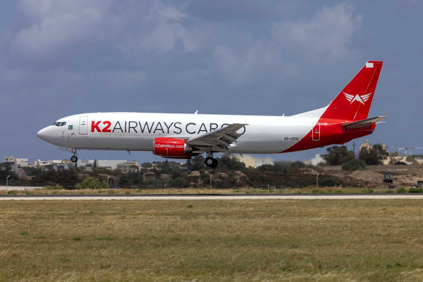 A side profile shot of a K2 Airways Cargo Boeing 737 freighter just moments before touching down on a runway. The aircraft features a clean white fuselage, bright red engine nacelles, a red tail section with a white winged emblem, and the registration code VP-CFG visible near the rear.