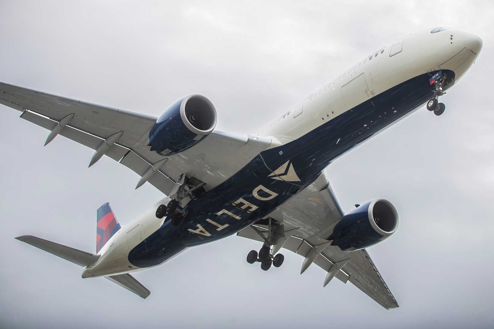 Delta Air Lines A350-900 captured from below, showcasing the aircraft's underbelly, engines, and wing detail against a cloudy sky