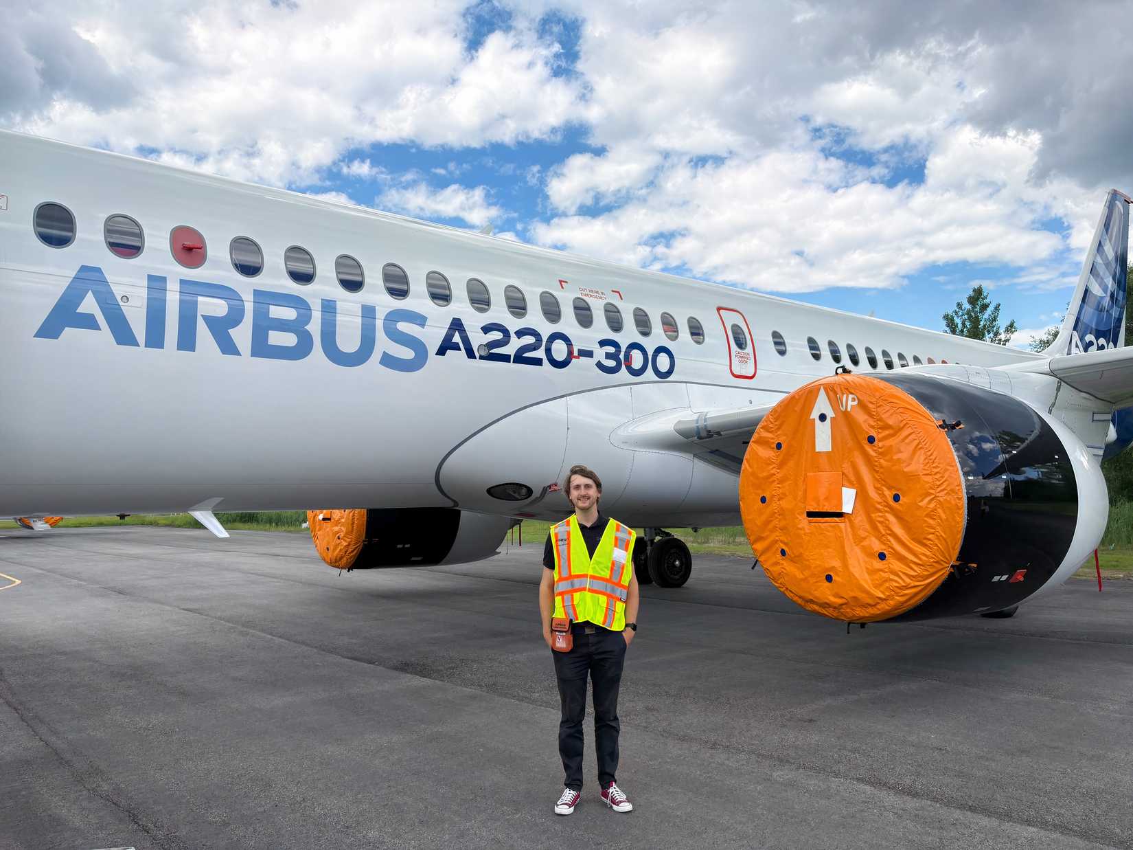 A full-length portrait of Tom Boon wearing a high-visibility safety vest standing on an outdoor tarmac directly in front of an Airbus A220-300 test aircraft, which features prominent blue "AIRBUS A220-300" branding and bright orange protective covers over its jet engines.