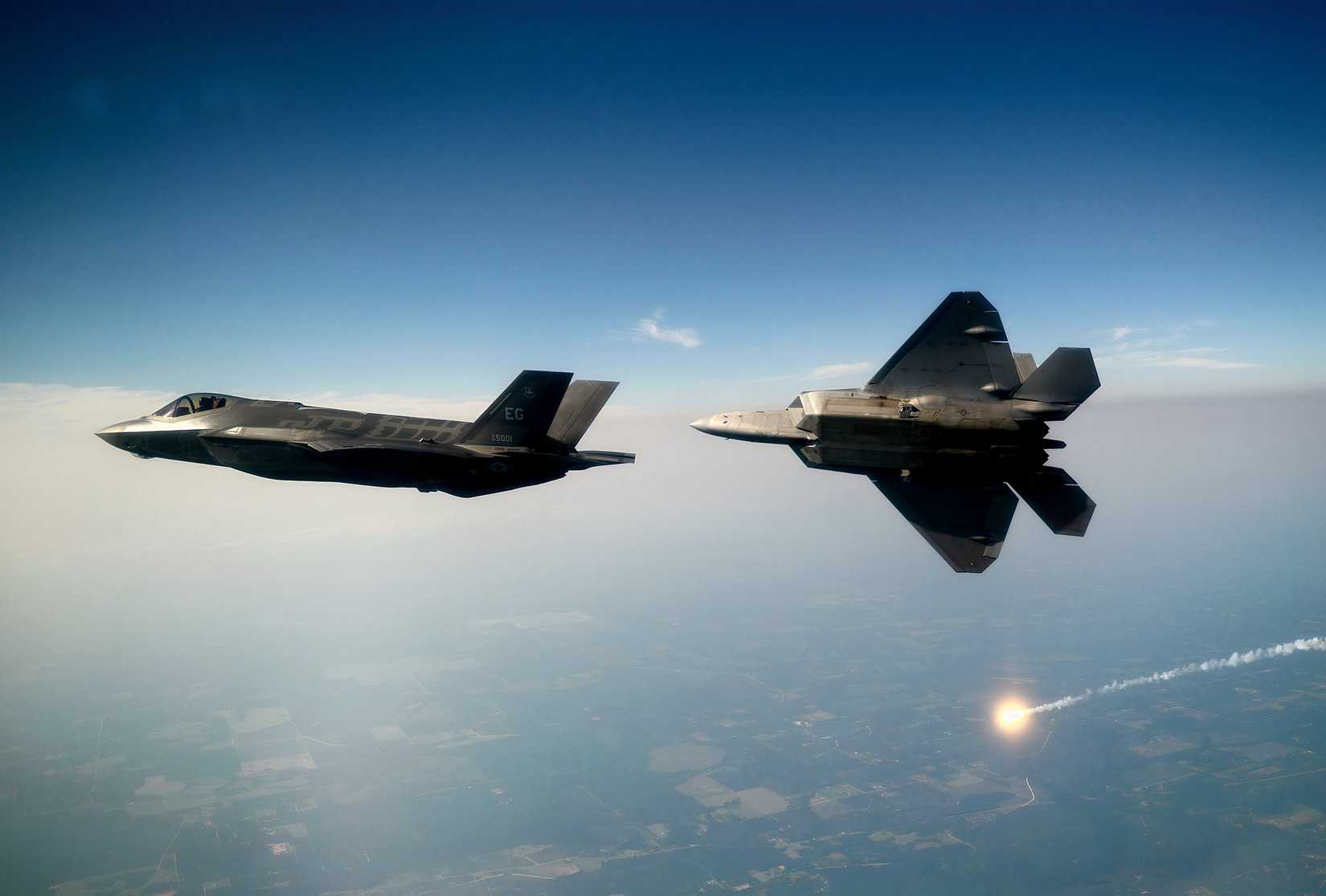 A photo of two F-35s, flying high above a rural area, one of the aircraft is deploying a flare while maneuvering away from the other. 