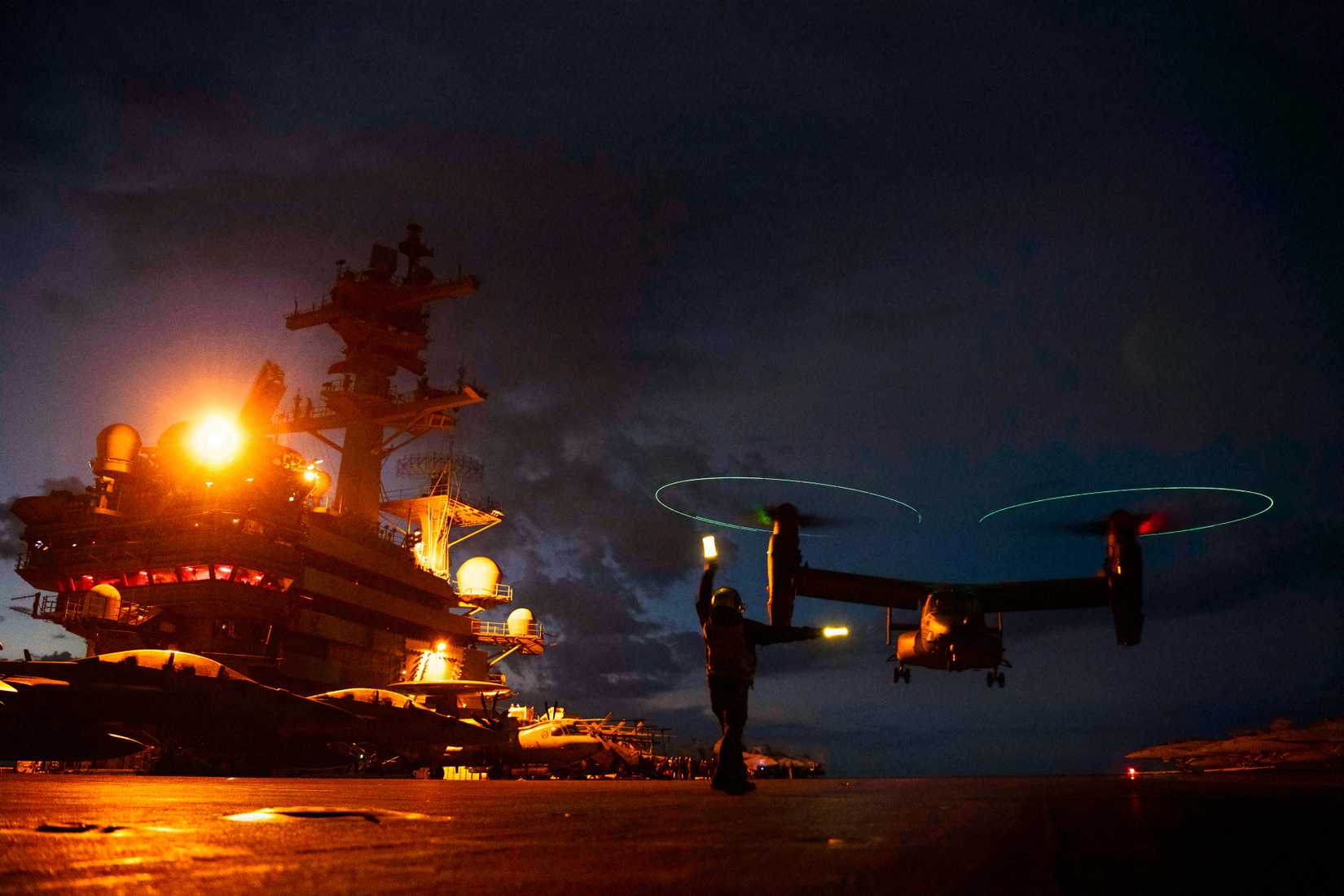A photo of an Osprey making a night landing on a carrier.