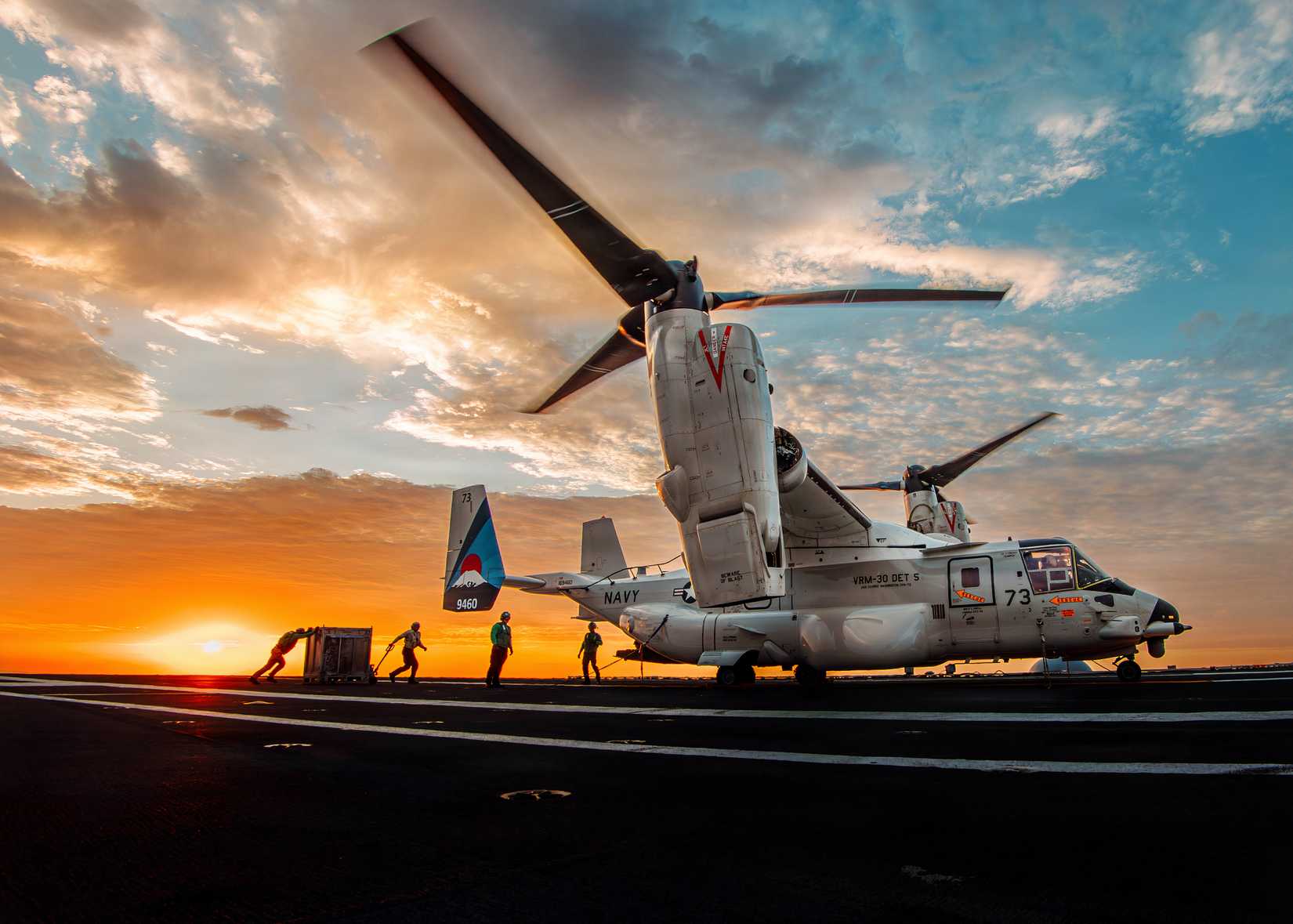 A photo of an Osprey that has just landed on a carrier. The background shows a sunset with crew rushing to move cargo to the aircraft. 
