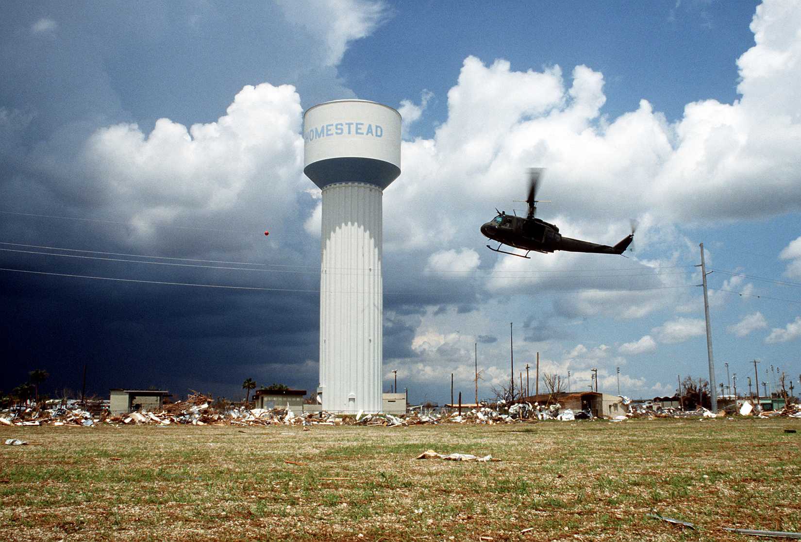 A UH-1 Iroquois helicopter hovers over the destruction caused by Hurricane Andrew while a water tower remains untouched by the storm