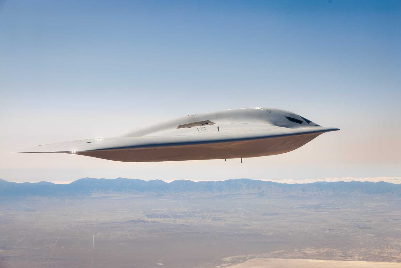 A photo of a B-21 flying over an arid background, against a hazy blue sky.