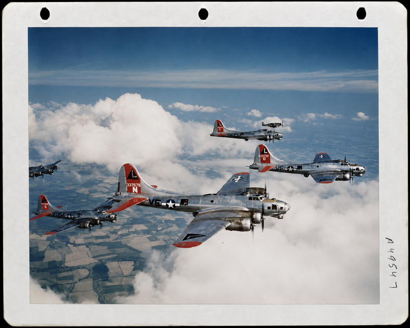 A photo showing a group of B-17s being escorted by a P-51 in the background. The photo was likely taken over the English countryside or Germany.