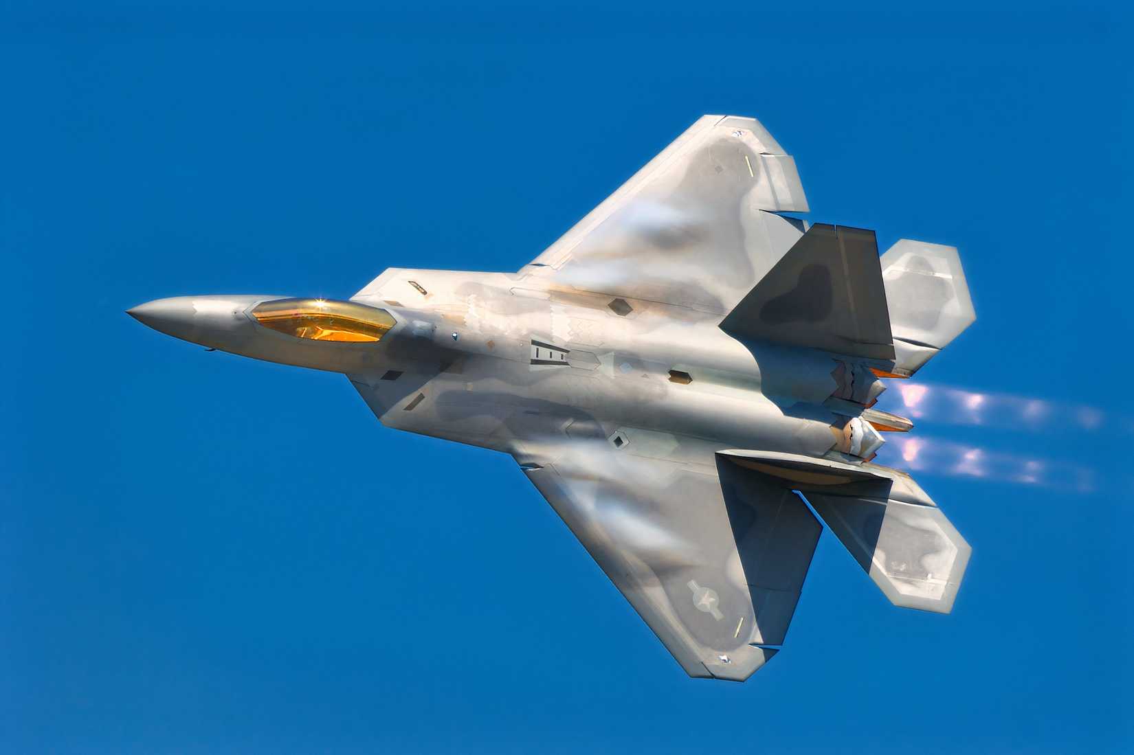 A photograph of an F-22 flying with the top of the aircraft toward the viewer, against cloudless blue sky. 