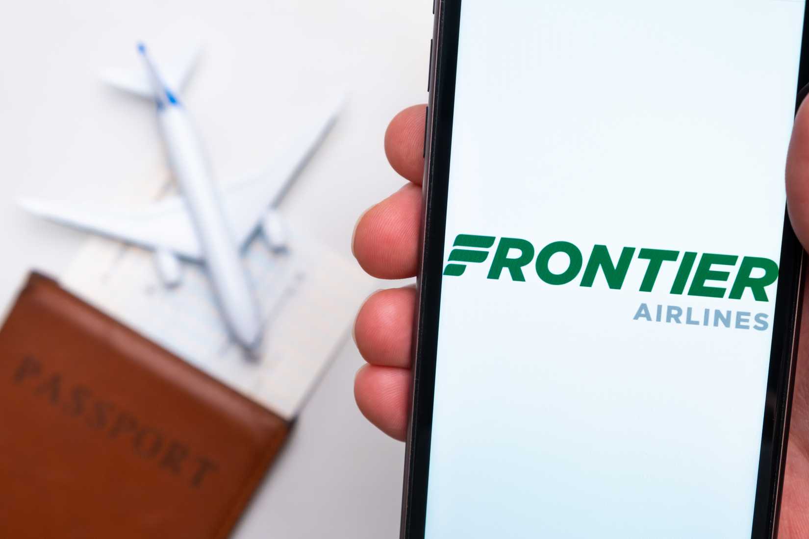 Frontier Airlines Airline application is displayed on the smartphone screen. There is a blurry plane, passport and boarding pass on the background. 