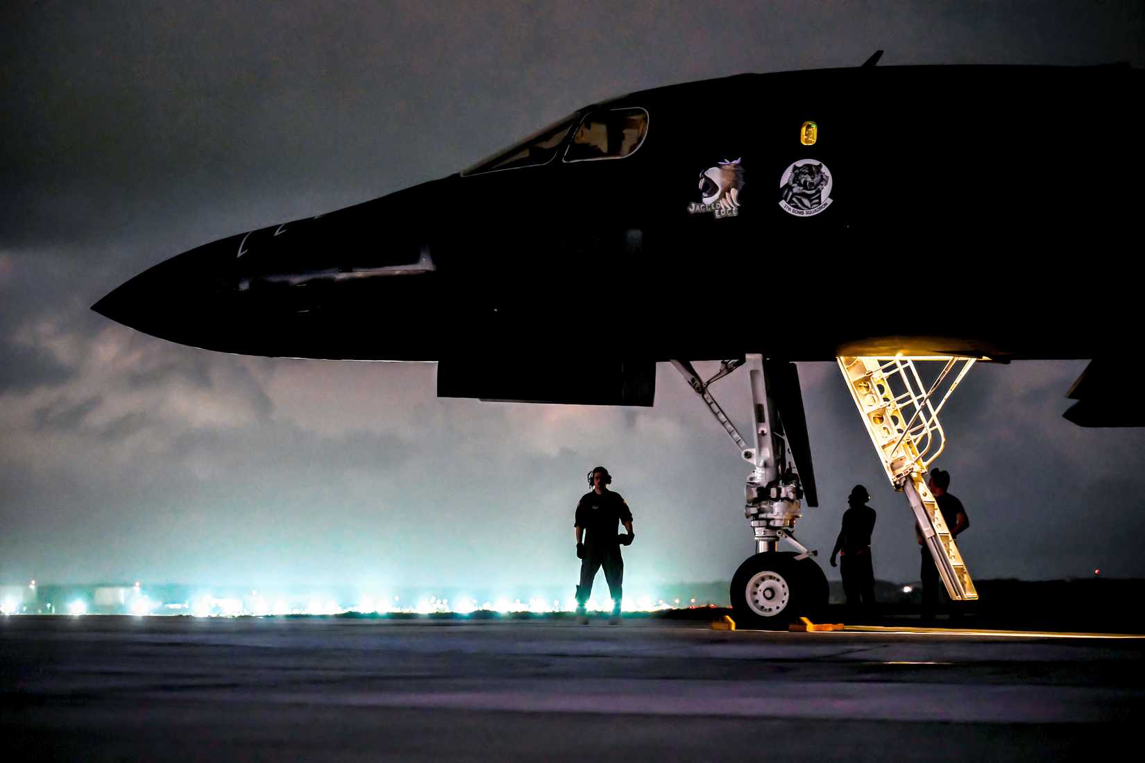 A close-up photo of the nose section of a B-1B at night, with lights in the background and a pilot or ground crew standing below the nose.