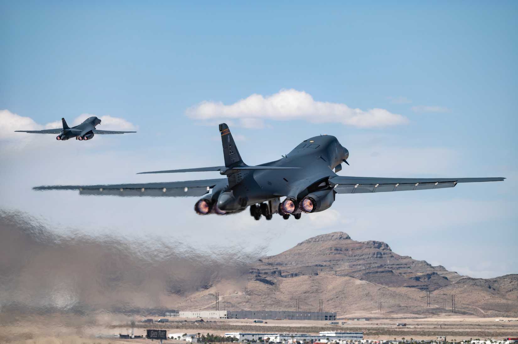 A photo of two B-1s just after taking off from their base with a arid mountainous scene in the foreground.