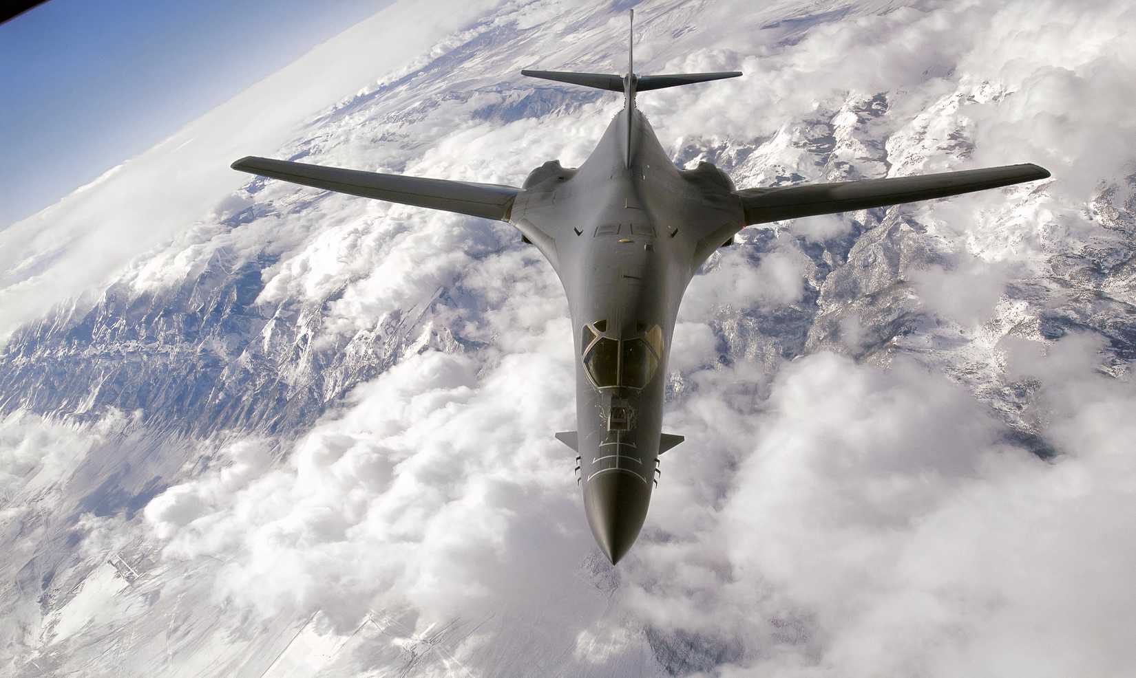 USA photo of a B-1 bomber flying over a cloud formation with mountains visible below.