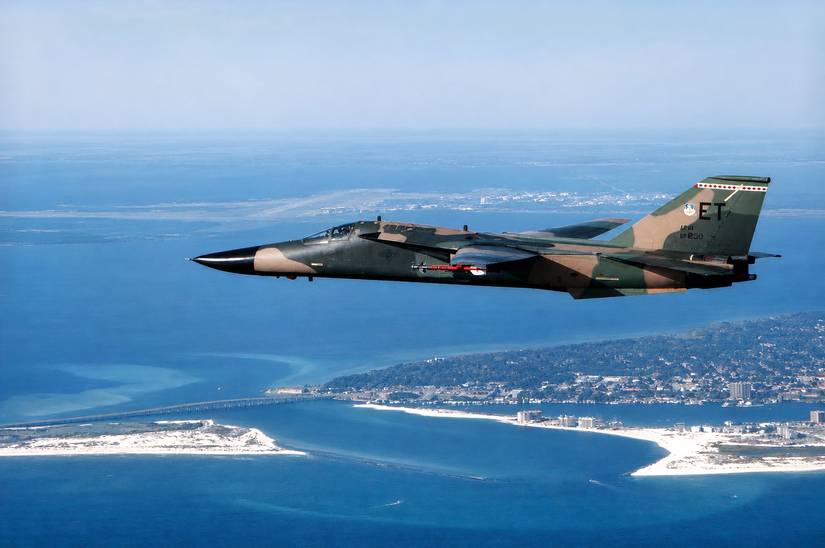 A photo of an F-111 Aardvark flying over water with a land features in the background.