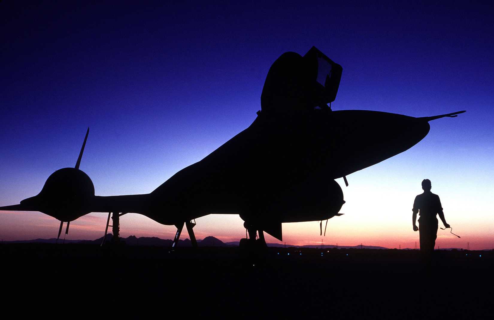 A technician carrying a twist drill stands beside an SR-71 aircraft silhouetted on the flight line