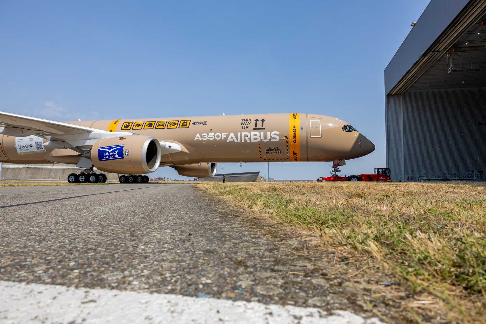 A wide side-profile shot from ground level of the Airbus A350F freighter prototype rolling out of an aircraft hangar attached to a red tug vehicle. The unpainted tan fuselage features cargo packaging icons, "THIS WAY UP" directional arrows, and bold "A350F AIRBUS" markings along the main body.