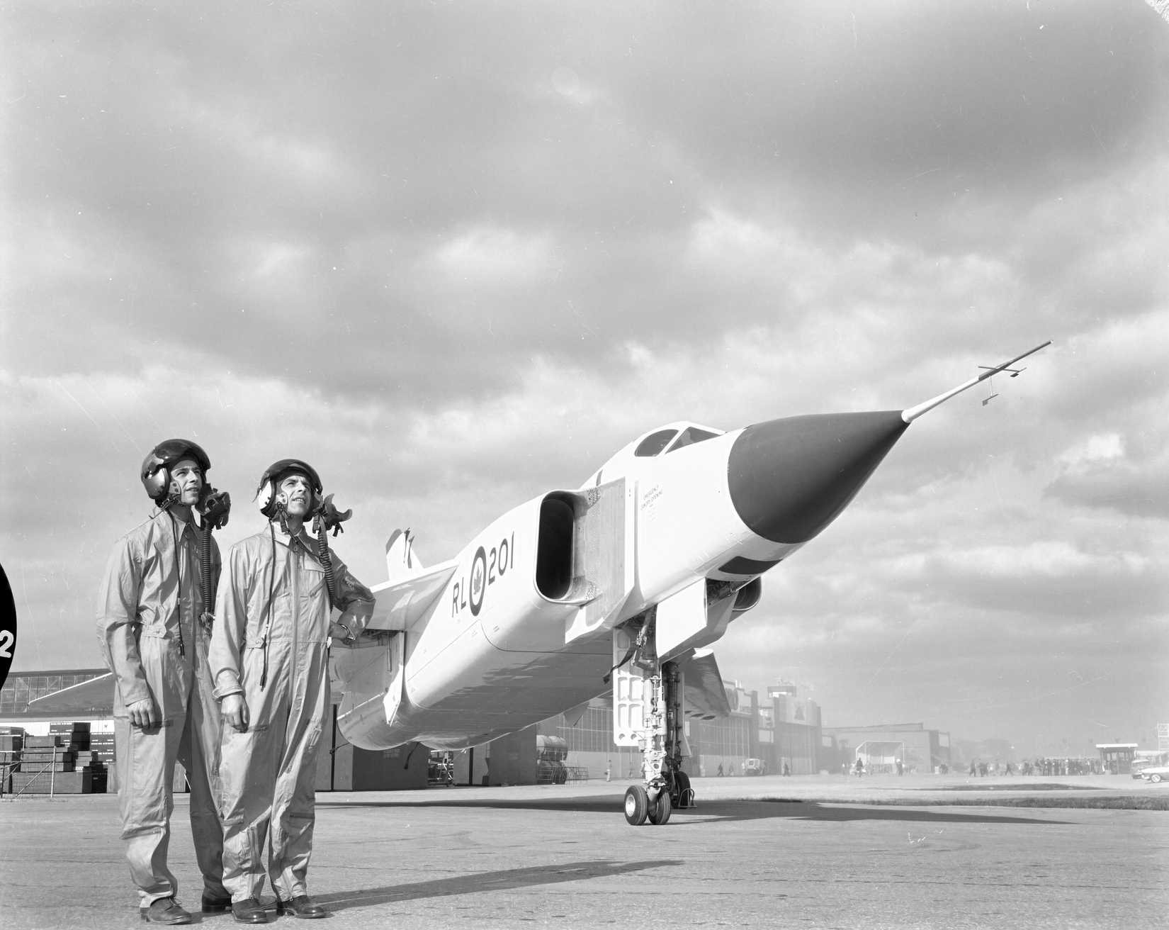 A black and white of the Avro Arrow parked on the tarmac, and two pilots in the foreground to the left of the aircrafts nose.