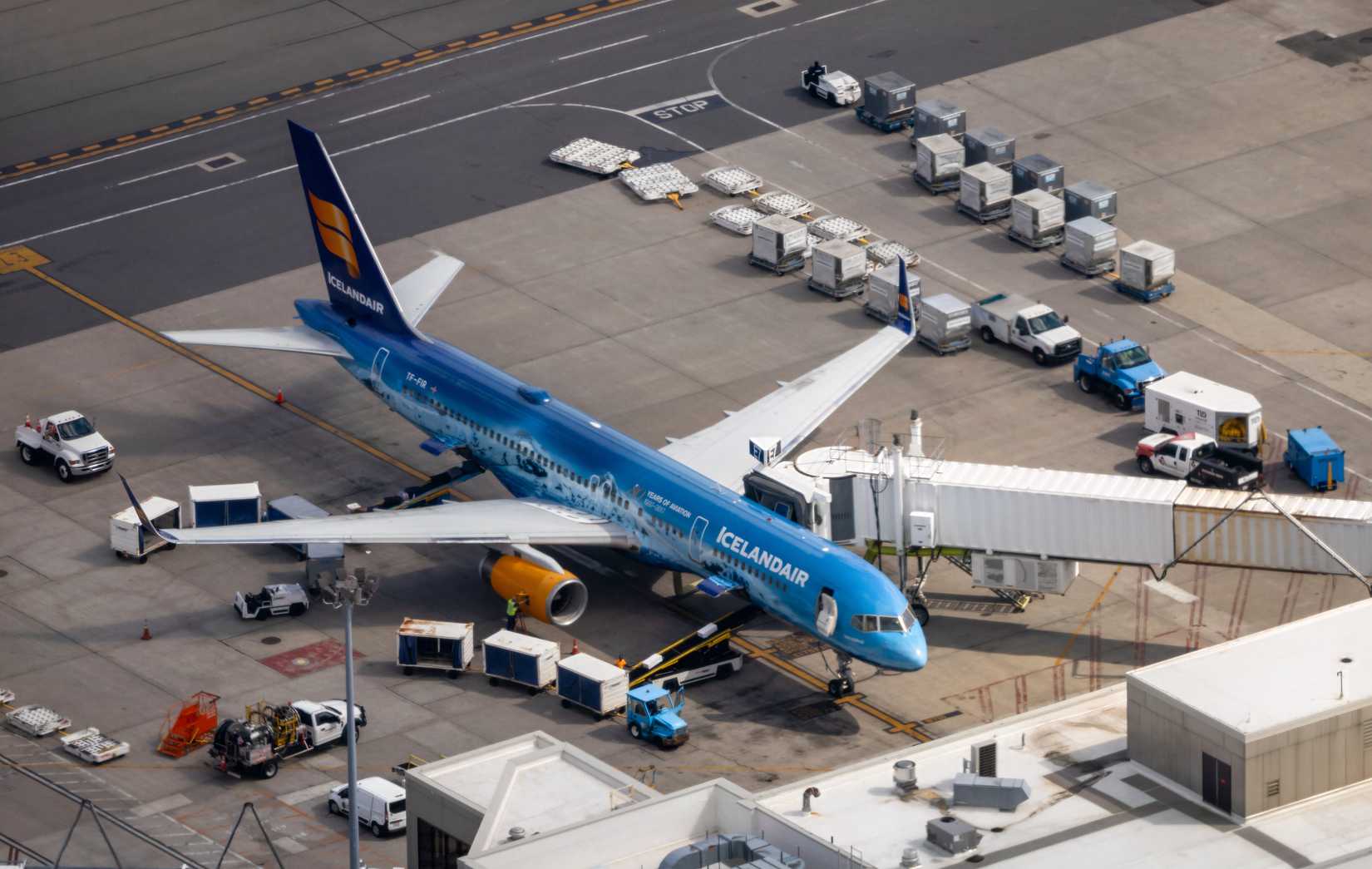 An aerial elevated view of an Icelandair Boeing 757 parked at an airport gate attached to a jet bridge. The aircraft features a special blue livery celebrating Icelandic aviation history, with yellow engine cowlings, surrounded by cargo containers, baggage tugs, and service vehicles on the tarmac.