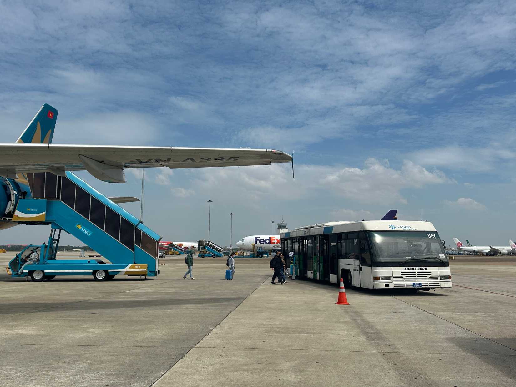 At Vietnam – February 26, 2025: "Passengers disembark from an aircraft at the airport tarmac, boarding a shuttle bus under a bright sky. Other planes and airport infrastructure are visible.