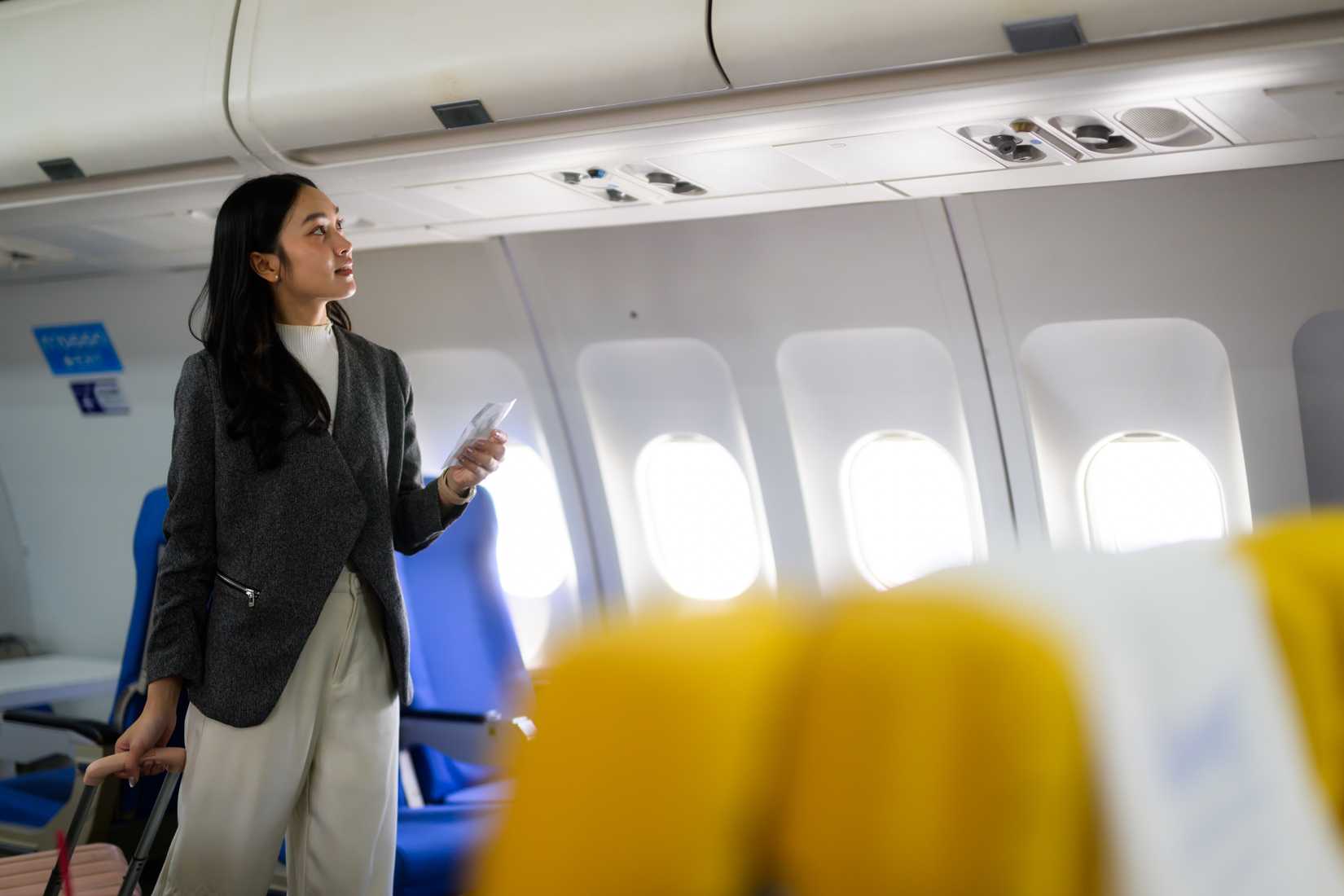 Young woman standing in airplane cabin holding a boarding pass with luggage, looking for a seat
