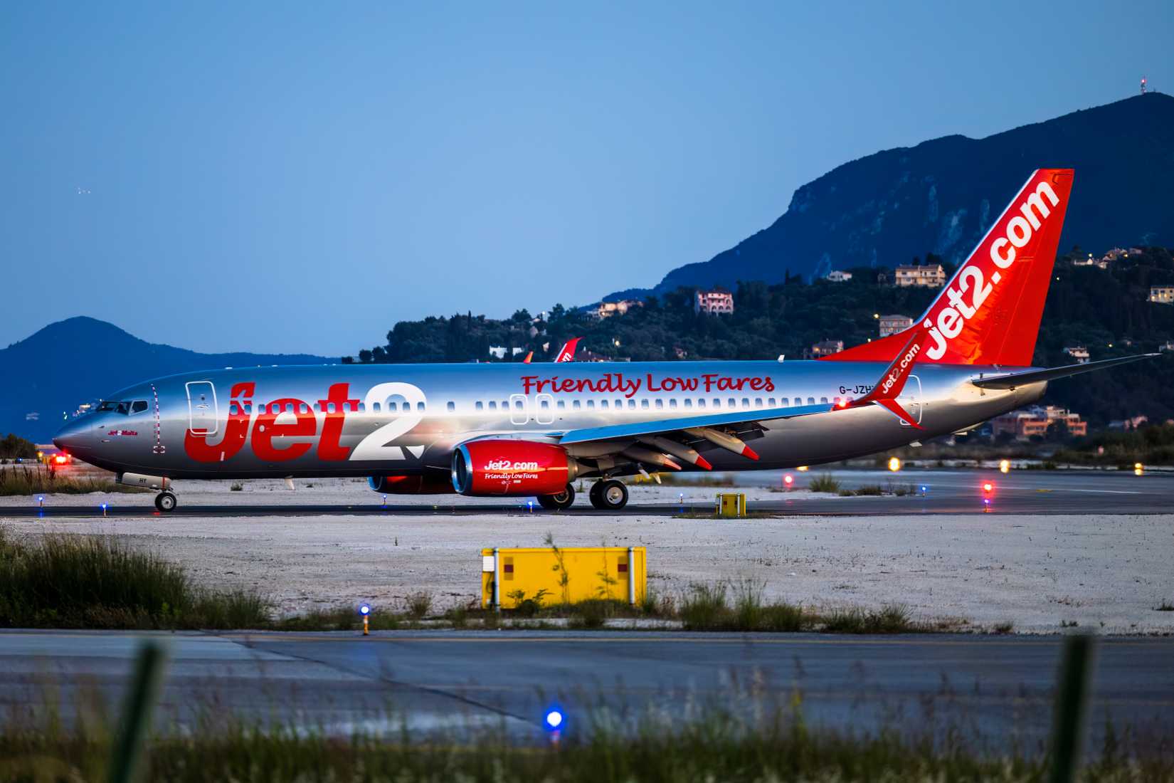 Greece Korfu Korfu 2025-06-06 A striking low-angle, evening shot of a Jet2 Boeing 737-800 (registration G-JZHY) taxiing on the runway at Corfu International Airport (Ioannis Kapodistrias) in Greece. The aircraft features Jet2's signature metallic silver fuselage with bold red "Jet2.com" branding and the slogan "Friendly Low Fares." The landing lights are brilliantly illuminated, casting a powerful glow across the runway, while the darkened, tree-covered hills of Corfu and a few scattered villa l