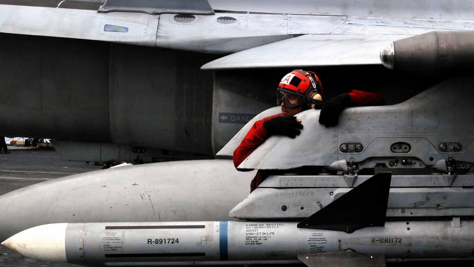 A photo showing the AIM-7 Sparrow mounted on an aircraft with an Navy ordnance technician clinging to the wing pylon above the missile.