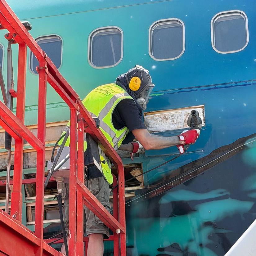 A worker standing on a red scaffolding platform while using a power tool to cut into the side of an aircraft fuselage. Wearing a high-visibility safety vest, ear protection, and a respirator mask, the worker is cutting panels from a retired airplane featuring a green northern lights livery below a row of cabin windows.