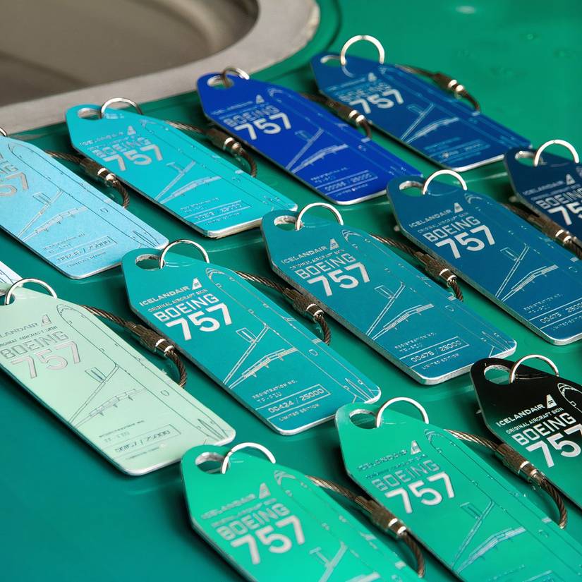 A high-angle, close-up shot showcasing an array of colourful Aviationtag keychains crafted from the skin of an Icelandair Boeing 757, registration TF-FIU. The tags feature printed aircraft schematics and unique edition numbers, arranged in rows across shades of mint green, teal, deep blue, and black.
