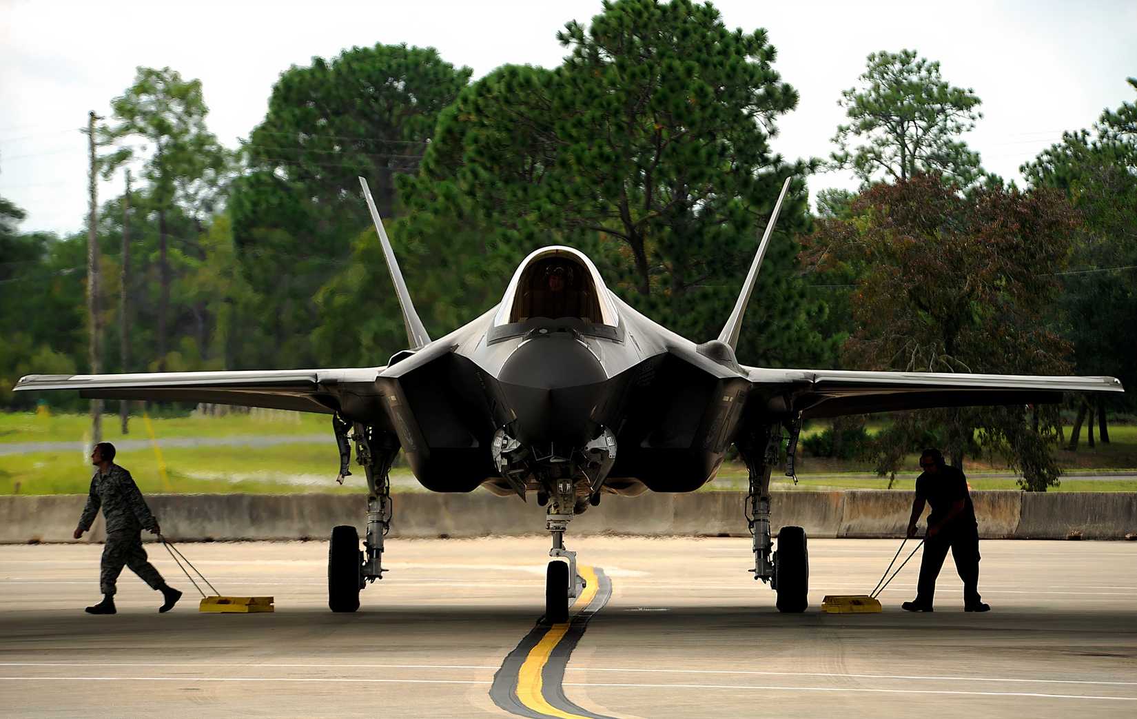 A photo of an F-35 having its stop-blocks removed from its wheels by ground crew, preparing to fly.