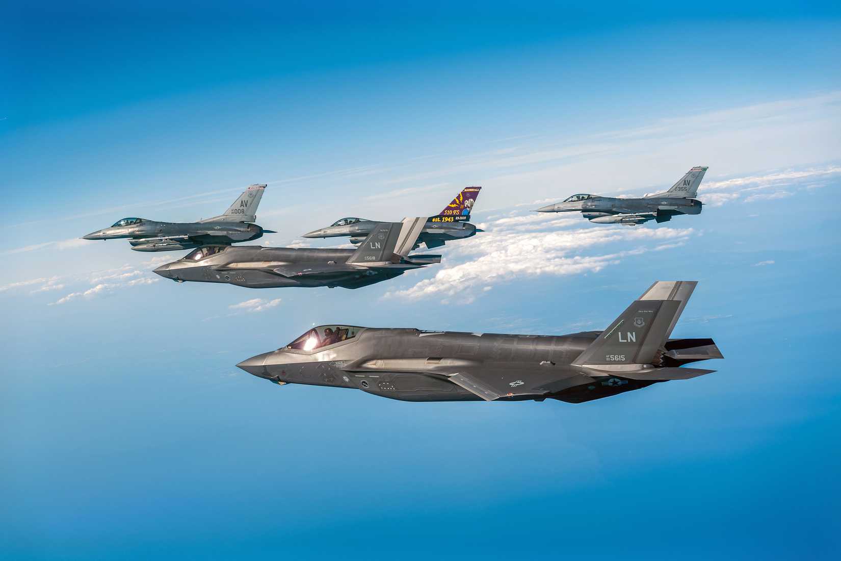 Two F-35s flying with three F-16s over a large body of water with clouds in the background.